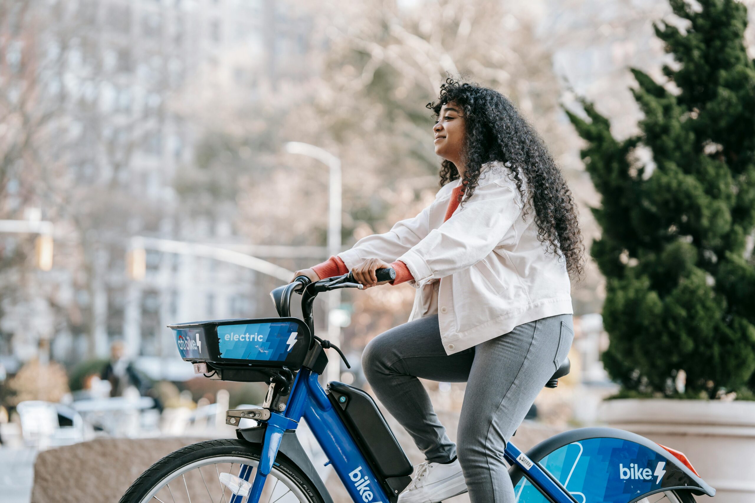 Side view of positive African American lady wearing casual clothes while riding bike in town in park in sunny day near buildings and trees